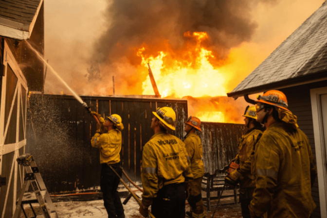 Firefighters hose down the roof of a home as flames and black smoke rise in the distance