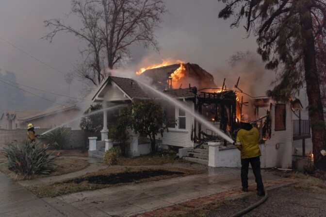 Two firefighters hose down a home that is nearly burned to the ground. The back half portion of the white home is destroyed and on fire