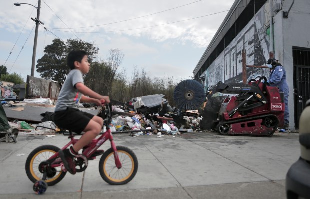 A young boy rides his bike along the sidewalk where Keep Oakland Beautiful and Clean workers, a division of Oakland Public Works department, removes a large trash pile along International Blvd. at 71st Avenue in Oakland, Calif., on Friday, Sept. 19, 2025. (Laura A. Oda/ Bay Area News Group)
