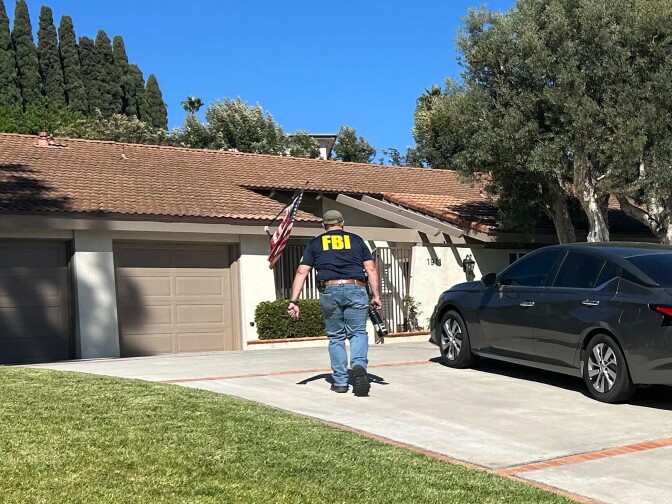 A man wearing a black shirt with yellow writing that reads "FBI" walks on the driveway towards a single story house with a large lawn and a car parked in the driveway. 