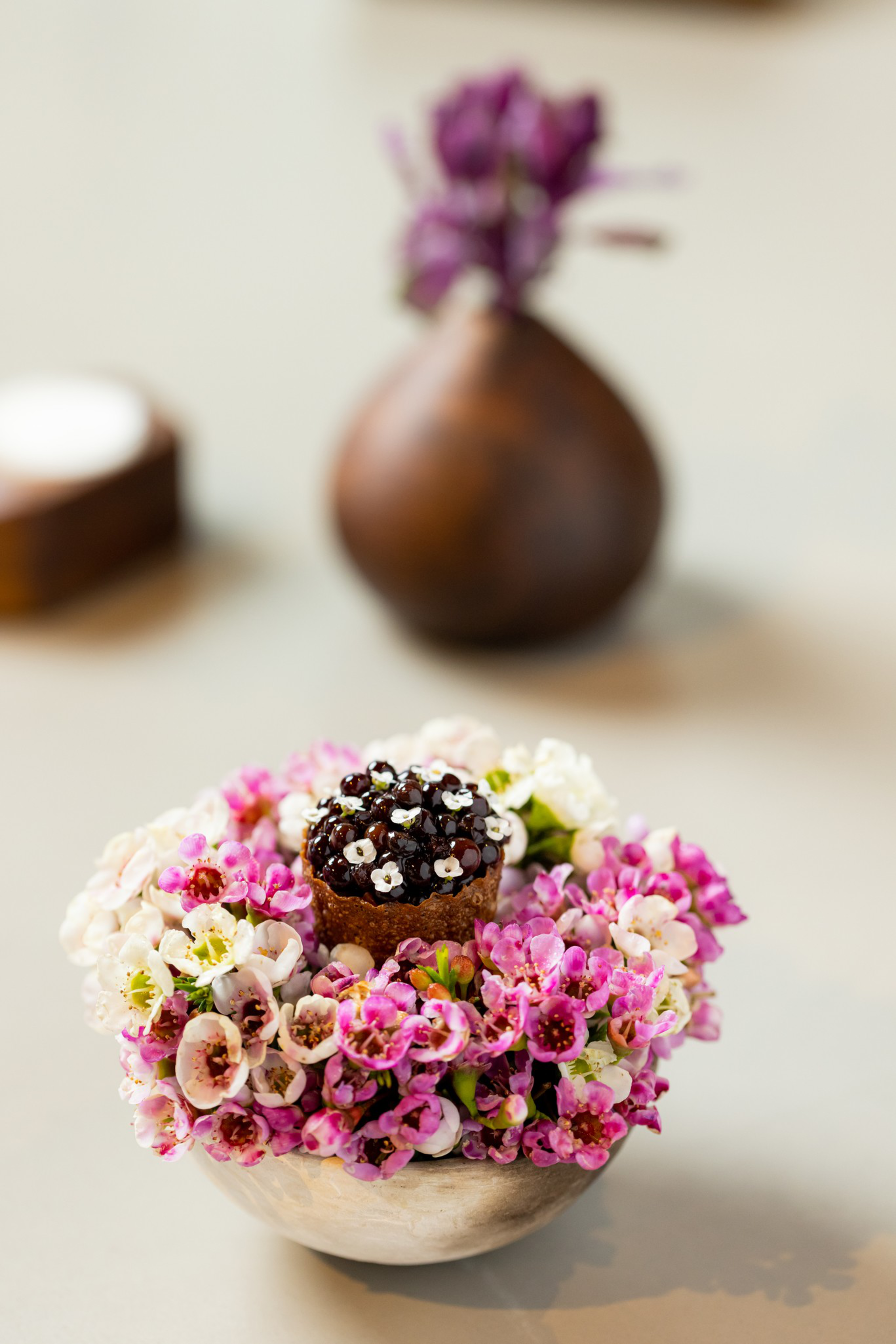 A small chocolate dessert topped with white flower petals rests in a bowl filled with pink and white flowers, with a blurred brown vase holding purple flowers in the background.