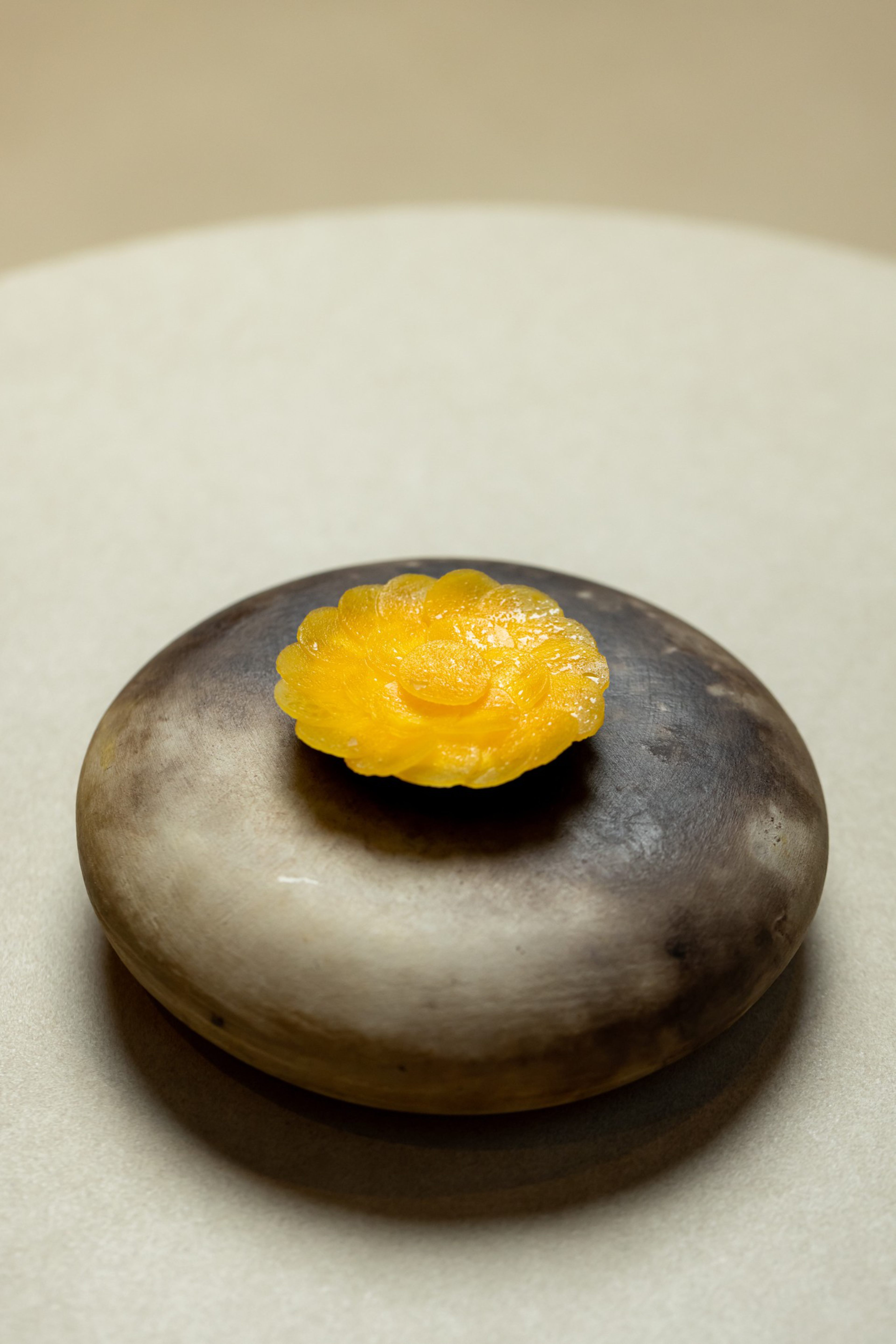 A small, yellow, flower-shaped gelatin dessert sits atop a smooth, round, dark-gray stone on a beige surface.