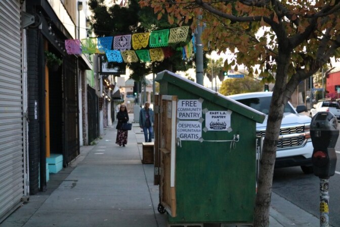 A green wooden storage is set on a sidewalk in front of shops right before the curb. The are signs taped to the side of the storage pantry that reads "Free community community" in English and Spanish.