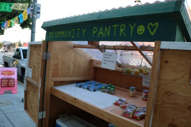 A wooden pantry storage on a sidewalk is open with canned and boxed items on its shelves. Oranges sit in a metal basket hanging from the top. A sign next to it reads in Spanish, "Take what you need, leave what you can." Painted on top in writing reads "Community pantry."