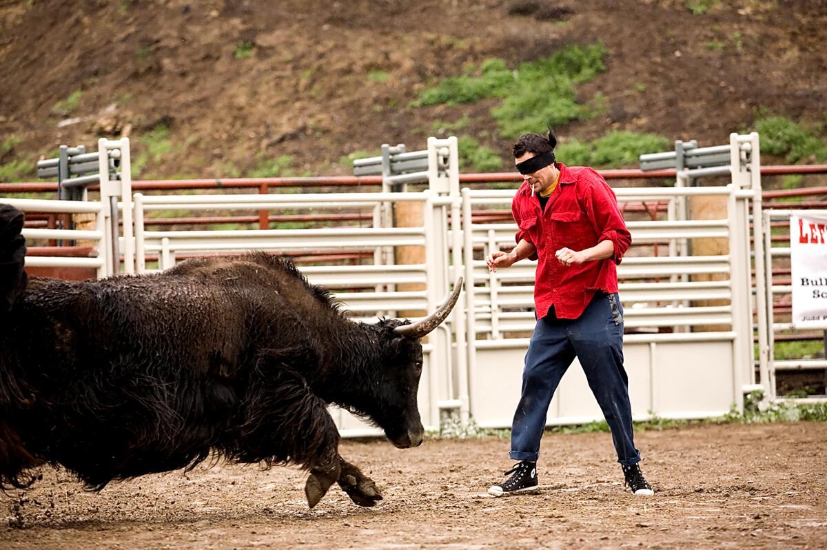 A blindfolded man awaits a charging bull.