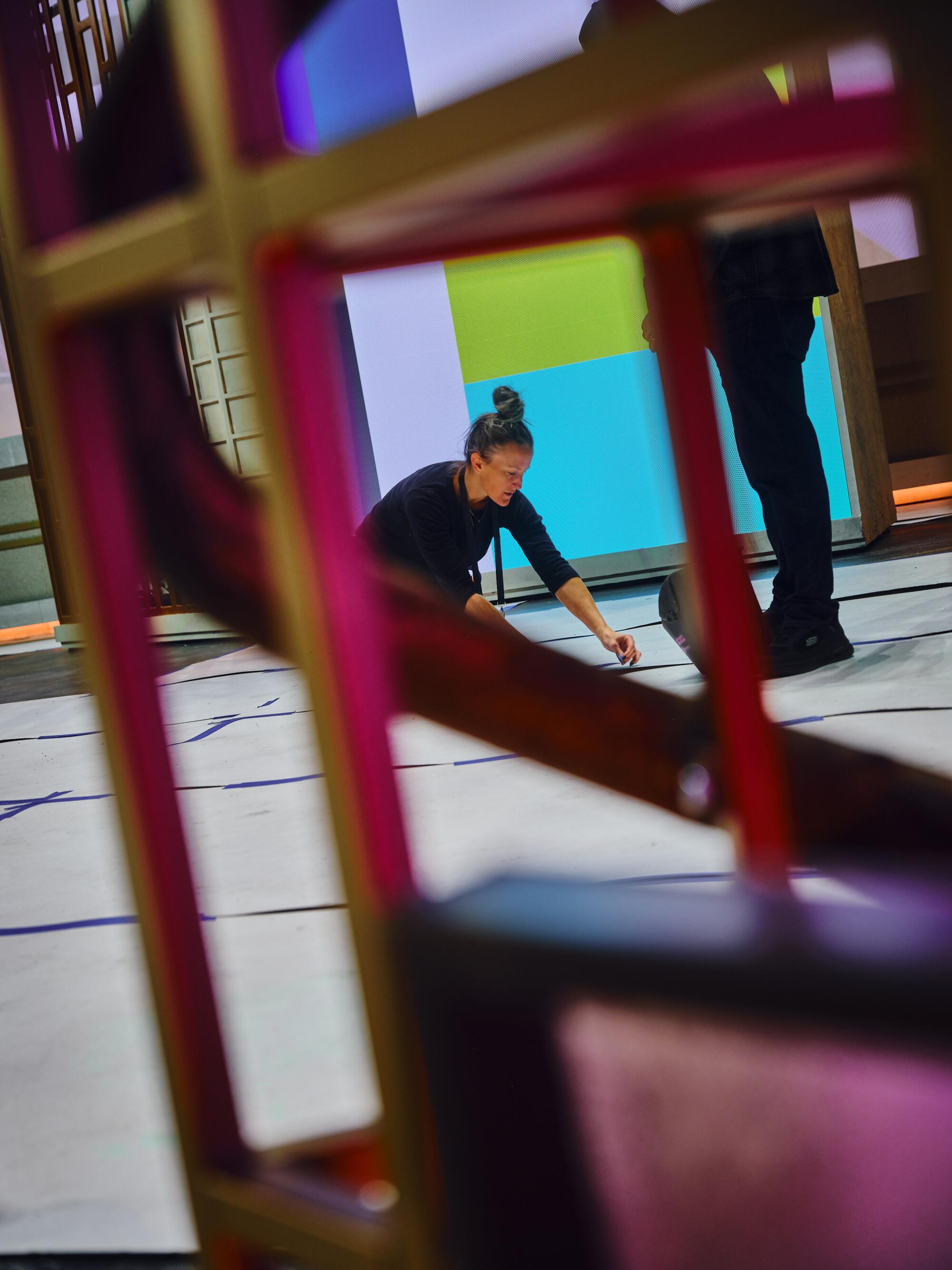 A woman sits on the ground, taping out a floor plan with white paper on stage. 