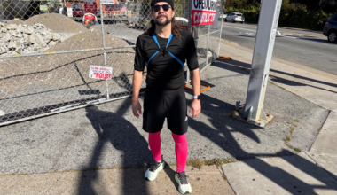 A man wearing a black outfit, pink leggings, and sunglasses stands on a sidewalk near a fenced construction site with debris behind him.