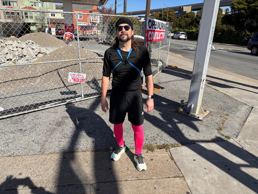 A man wearing a black outfit, pink leggings, and sunglasses stands on a sidewalk near a fenced construction site with debris behind him.