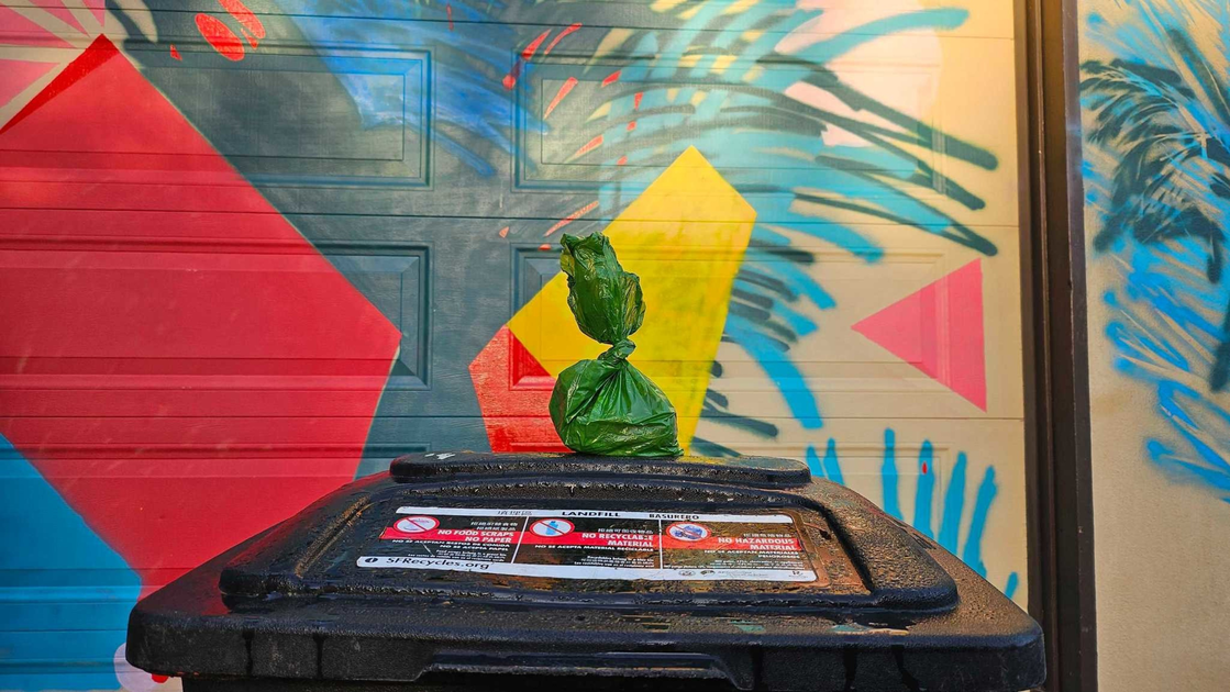 A small green plastic bag tied at the top sits on a black trash bin against a colorful geometric and abstract mural background.