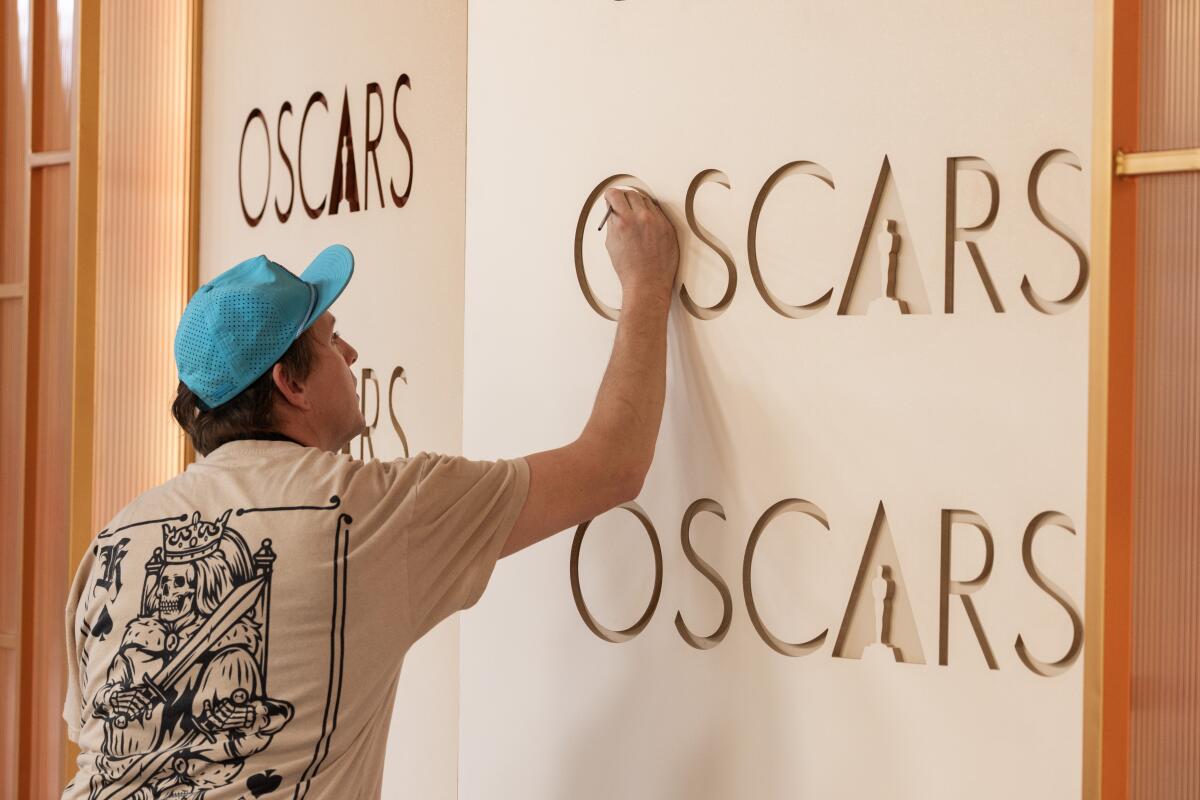 A man wearing a T-shirt and hat  touches up the red carpet backdrop for the Oscars