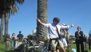 A man in a white shirt, tie, and cap stands with arms outstretched near a palm tree, surrounded by onlookers in a sunny park.