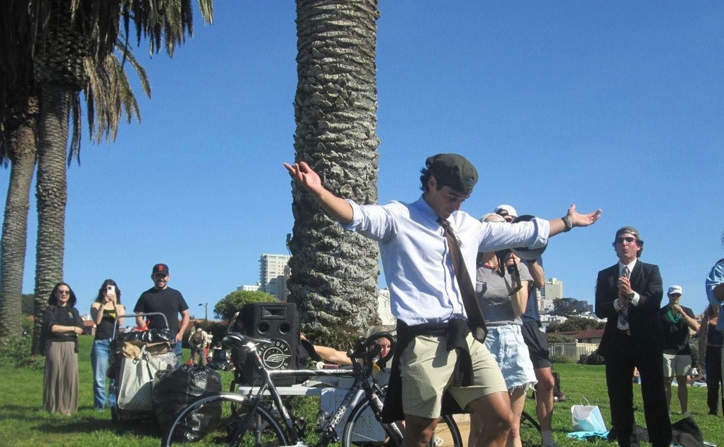 A man in a white shirt, tie, and cap stands with arms outstretched near a palm tree, surrounded by onlookers in a sunny park.