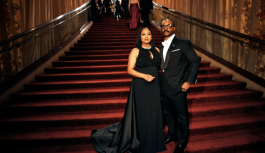 A man in a black tuxedo and a woman in a long black gown stand side by side at the bottom of a grand, carpeted staircase with gold handrails.