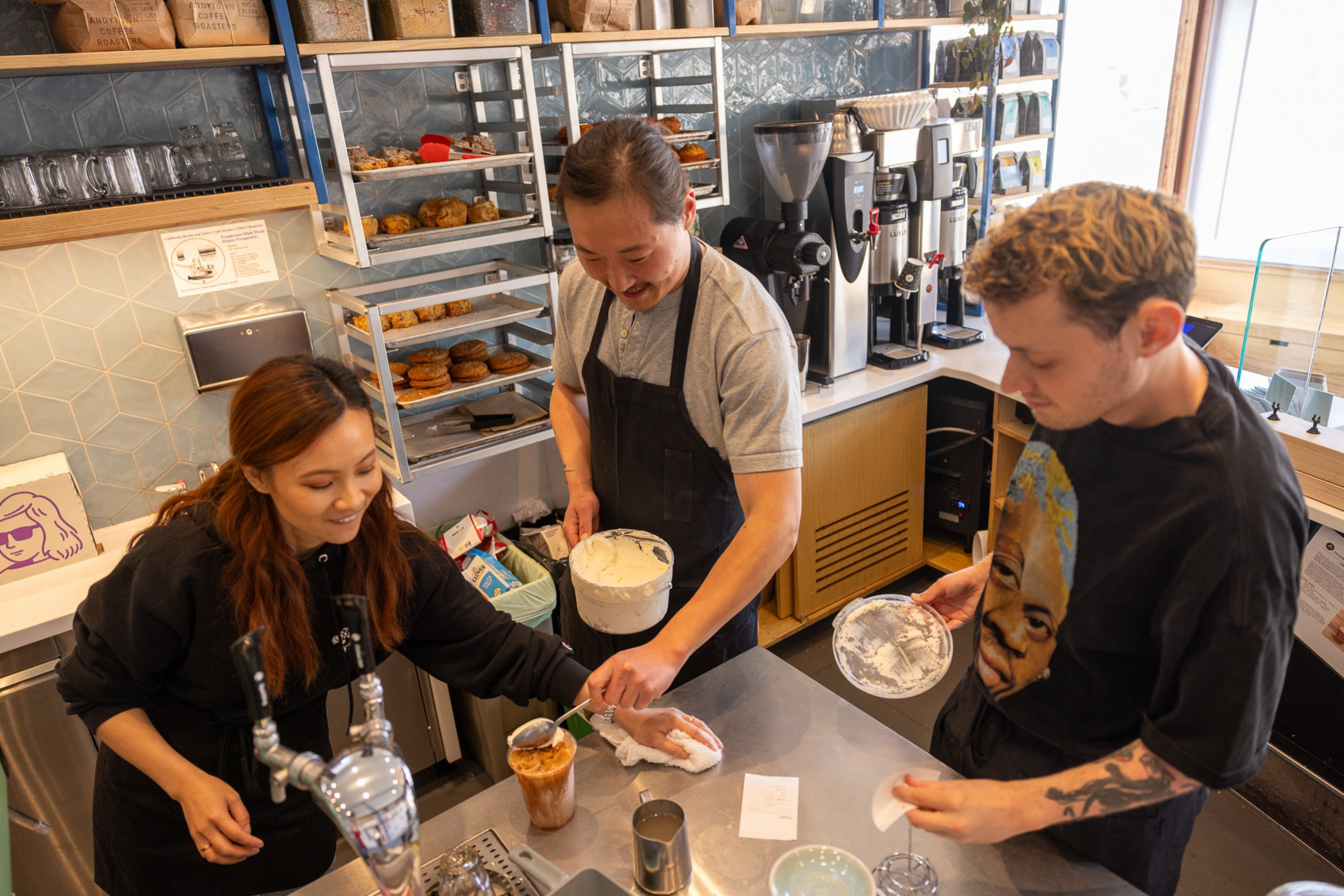 Three people are in a coffee shop, preparing drinks. They are smiling and working together. Shelves with pastries and coffee equipment are visible behind them.