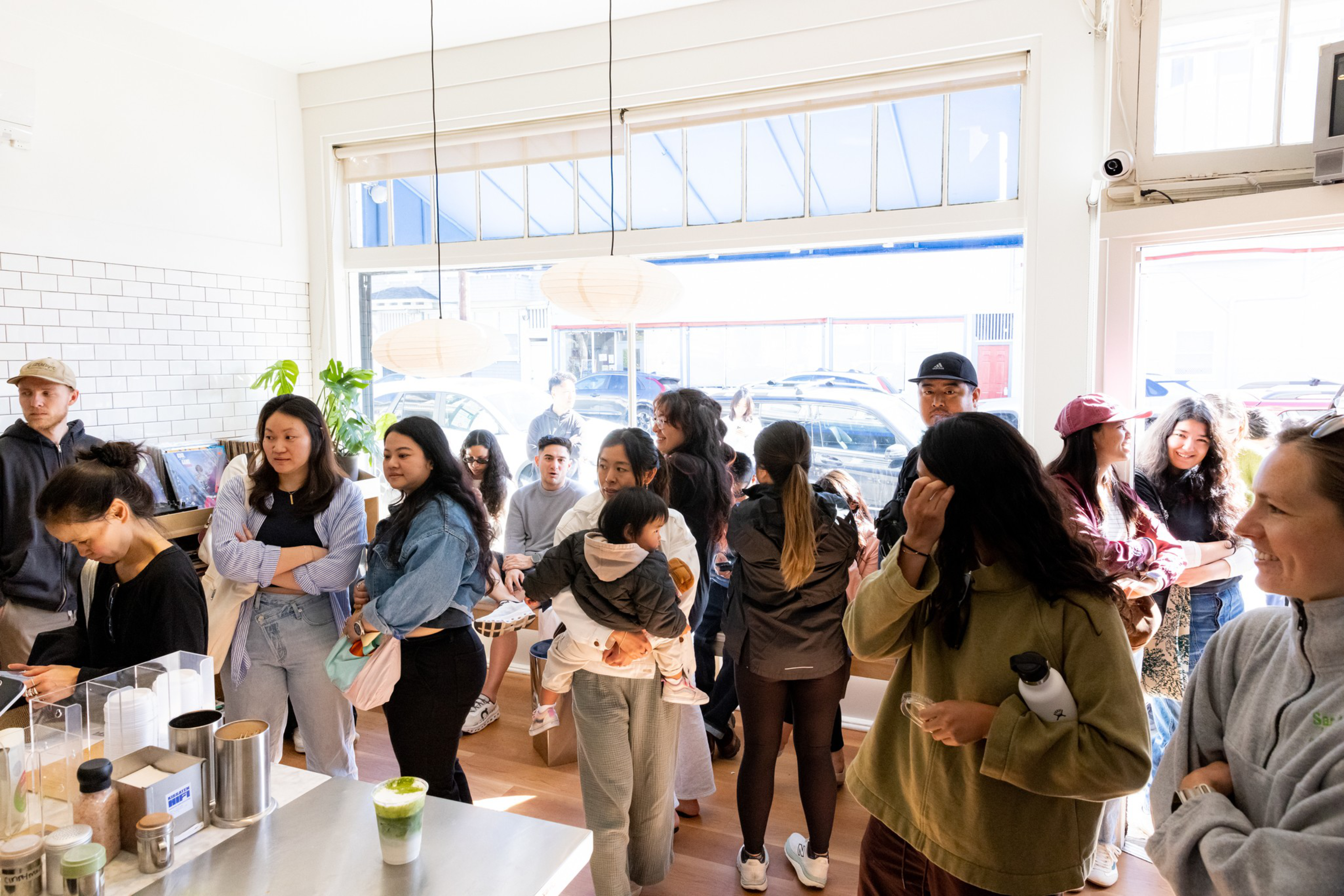 A group of people, including a child, wait inside a bright cafe near the counter and windows, some holding drinks and chatting.