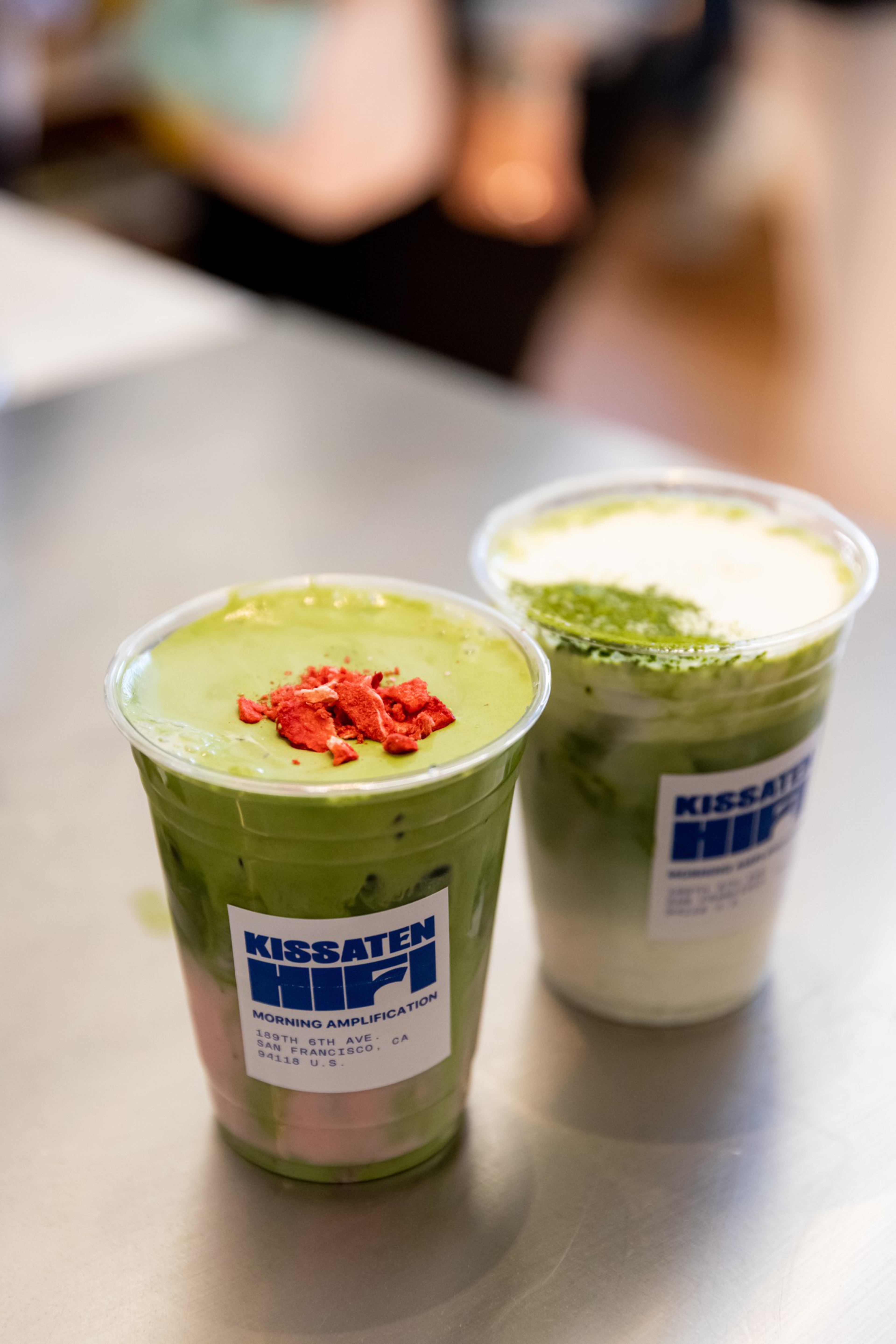 Two iced matcha drinks in clear plastic cups have layered green and white colors; one topped with red flakes and the other with matcha powder, labeled “KISSATEN.”