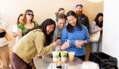 Three women laugh and take photos of drinks on a table, while several people stand and watch in the background.