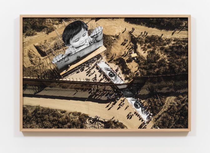 An aerial shot of an art piece depicting a large image of a child looking over a border fence.