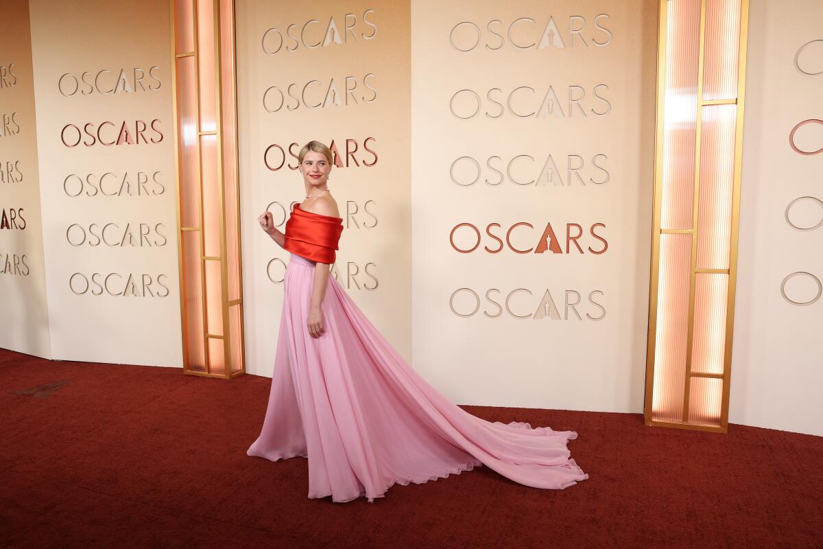 An actor in a pink dress and red top walks the red carpet.