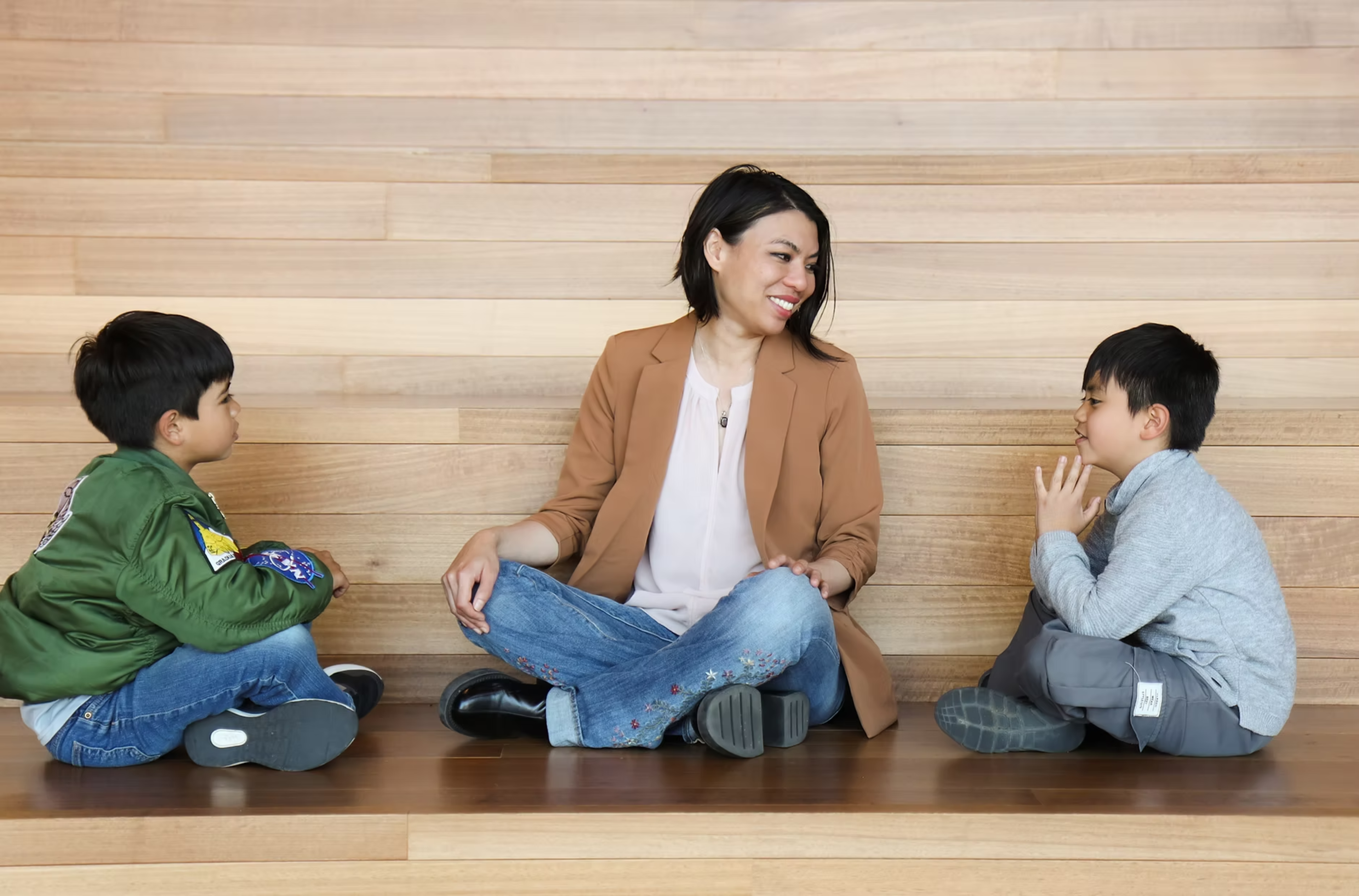 A woman sits cross-legged between two young boys, smiling and engaging with them while all sit on a wooden surface against a wood-paneled wall.