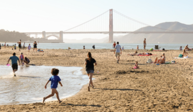 People relax on a sunny beach with the Golden Gate Bridge in the background. Children play near a shallow water area. Some are sunbathing on the sand.
