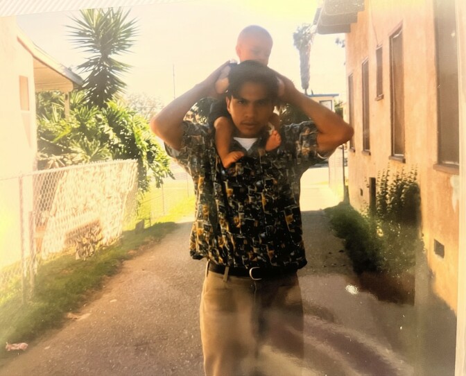 A young man with medium skin tone, short dark hair and a light mustache holds a baby over his shoulders. The father and son are in the driveway of a sun-bathed home.