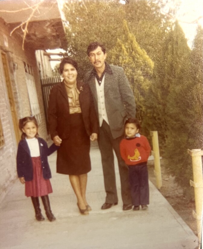 A family of four people with medium skin tone and dark hair — including a mother, a father, a small boy, and a little girl—pose for a photograph outside of a brick dwelling. The mother and daughter, both clad in long skirts, smile. The father and son are clad in pants and collared shirts.