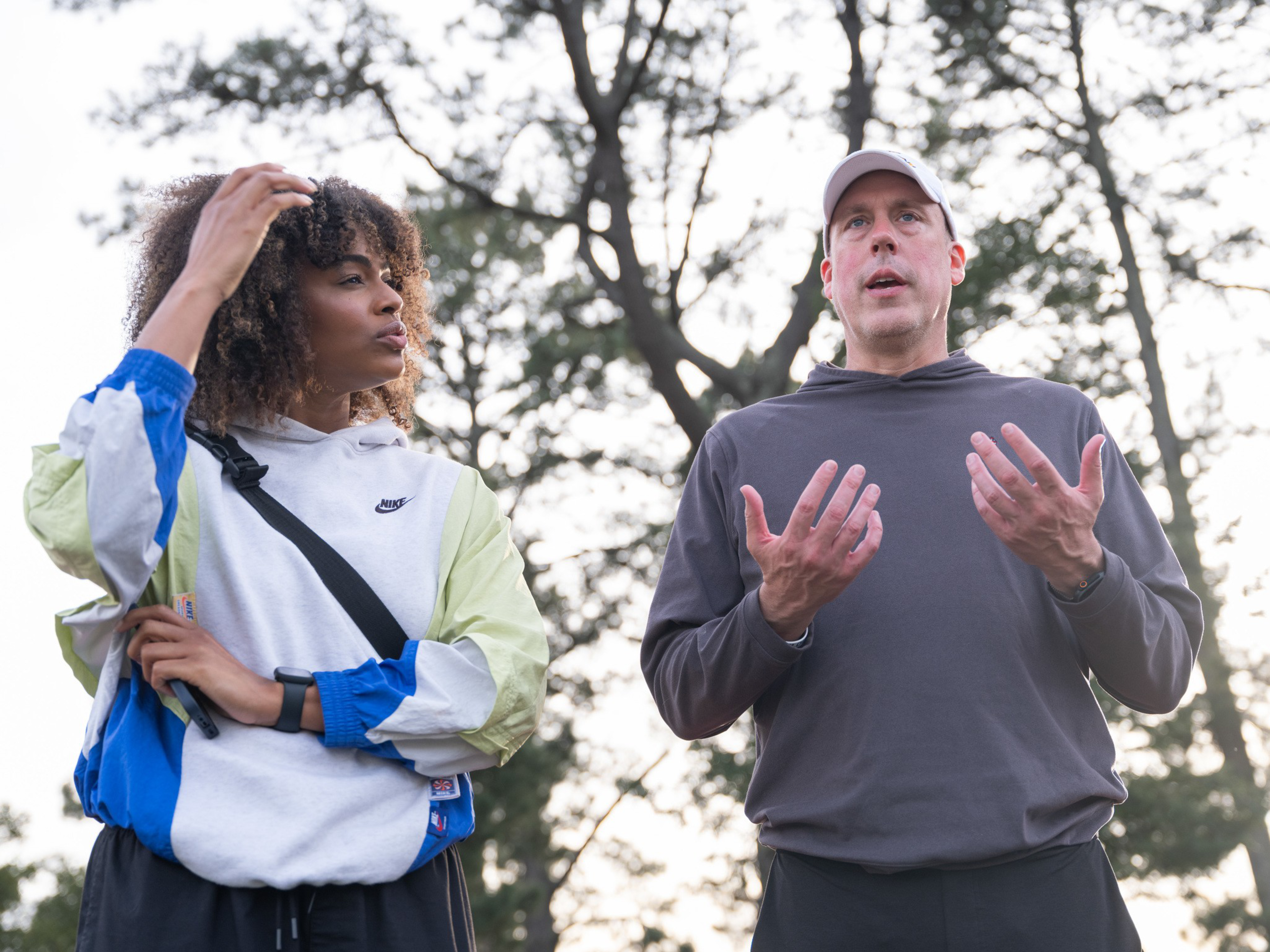 A woman with curly hair in a color-block Nike jacket talks while touching her head, and a man in a gray hoodie and white cap gestures with both hands.