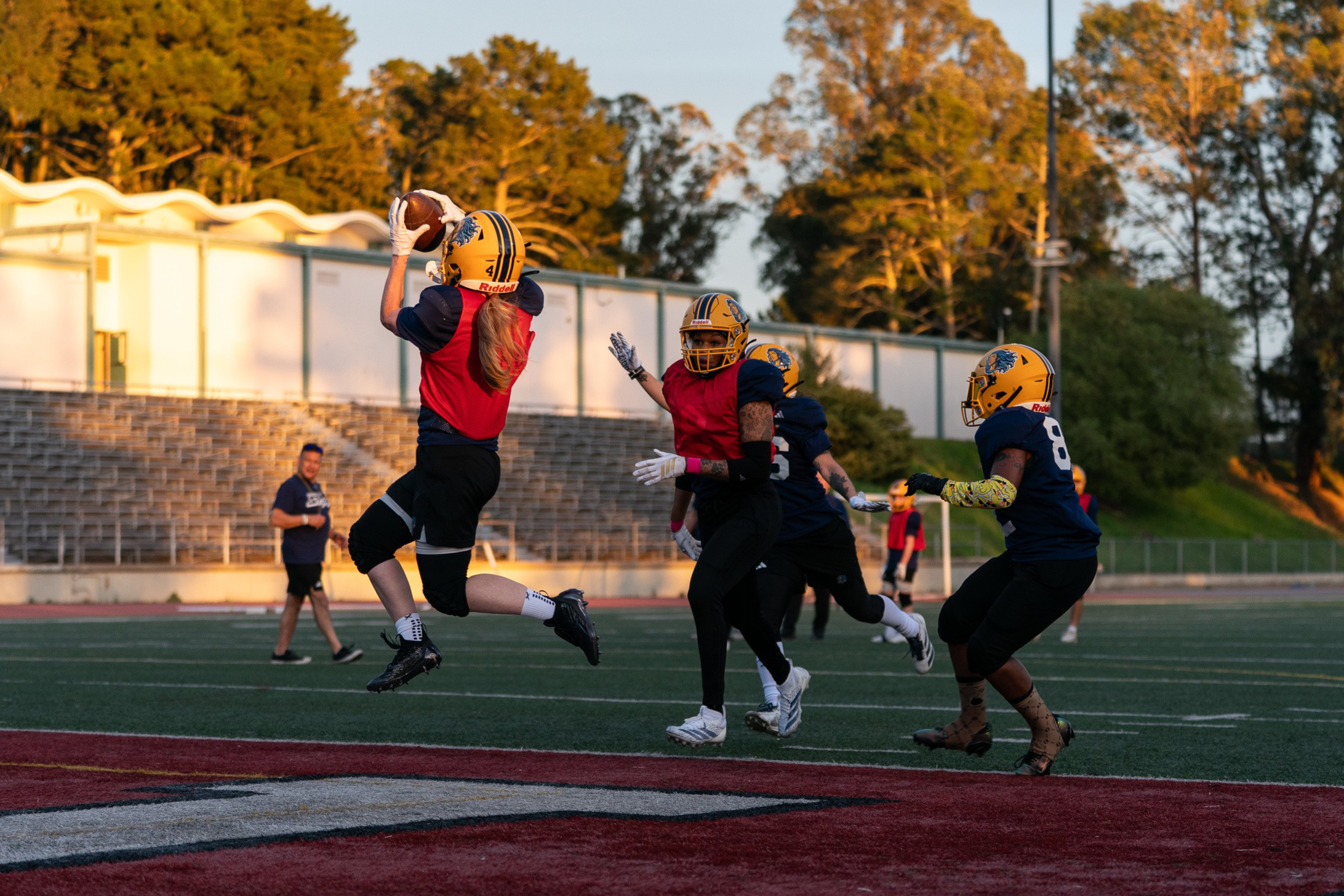 A football player in a red jersey and yellow helmet jumps to catch the ball while two teammates and an opponent run nearby on a field at sunset.