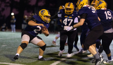A female football player in a yellow helmet and navy jersey runs with the ball while teammates block opponents on a lit field at night.