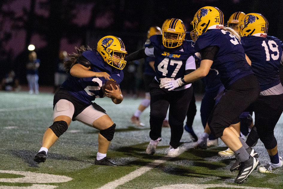 A female football player in a yellow helmet and navy jersey runs with the ball while teammates block opponents on a lit field at night.