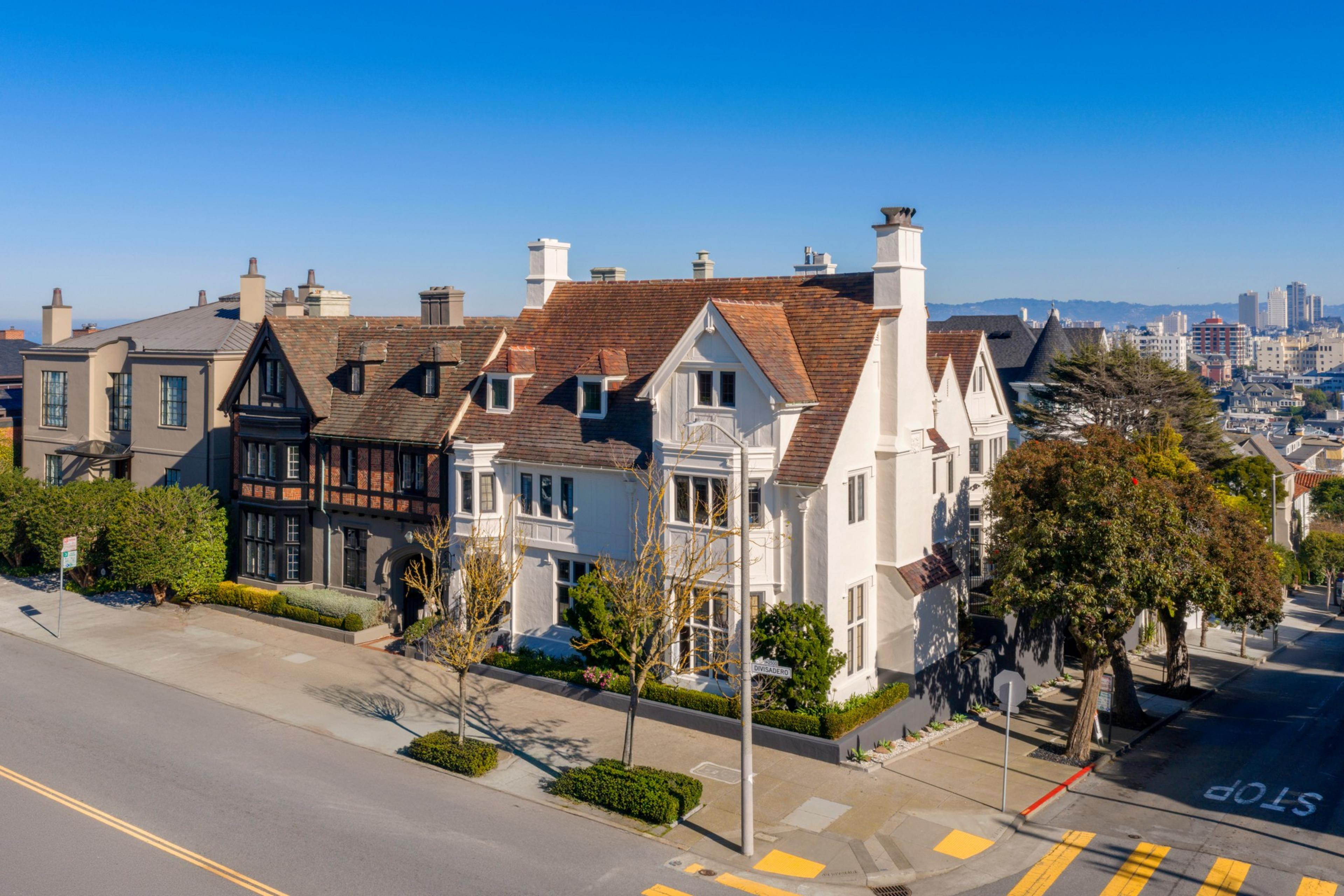 A street corner features large, well-maintained houses with varied architectural styles under a clear blue sky, with a city skyline in the background.
