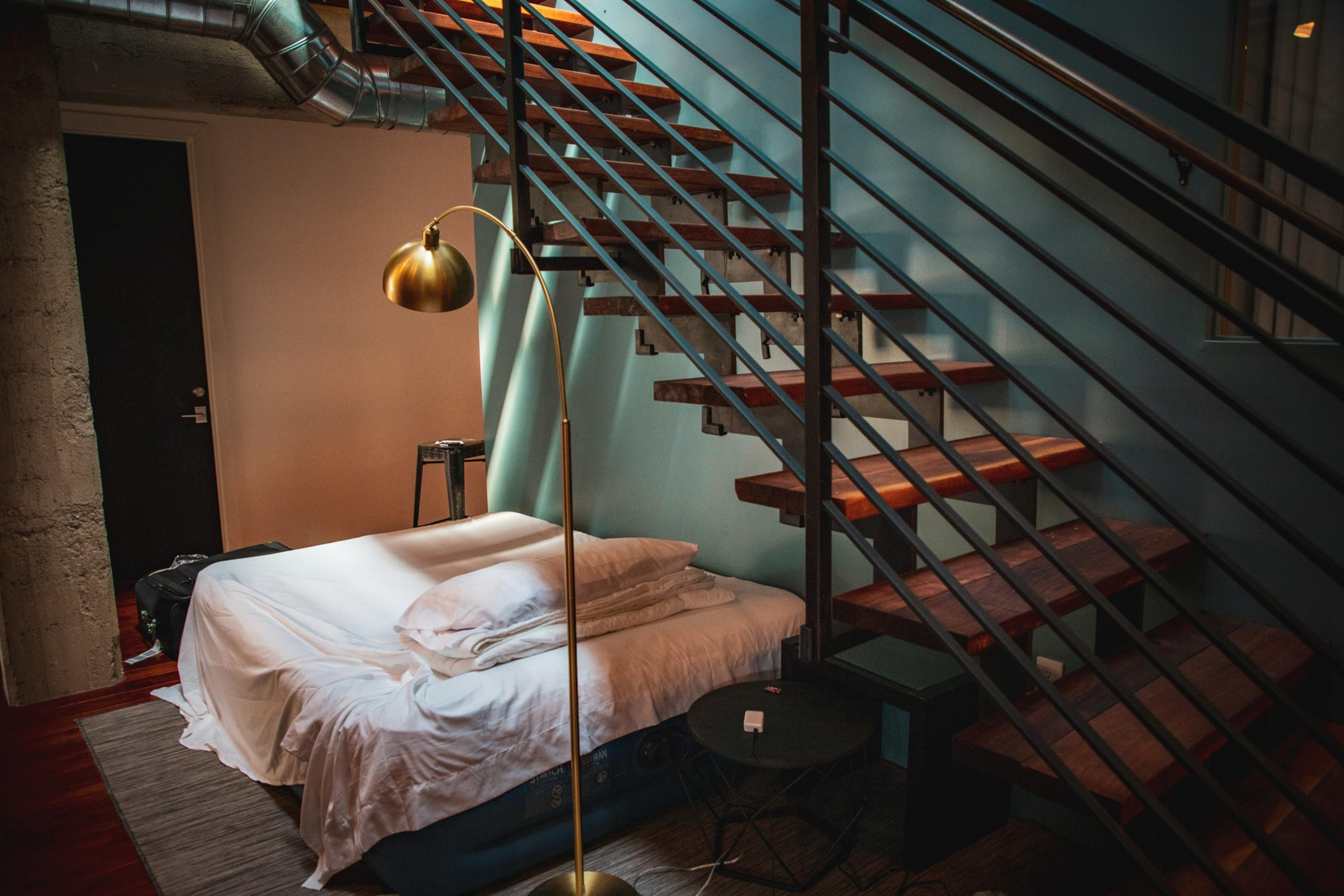 The image shows a loft-style room with a bed under wooden stairs, a gold floor lamp, and a small black table. The background includes exposed pipes and a door.