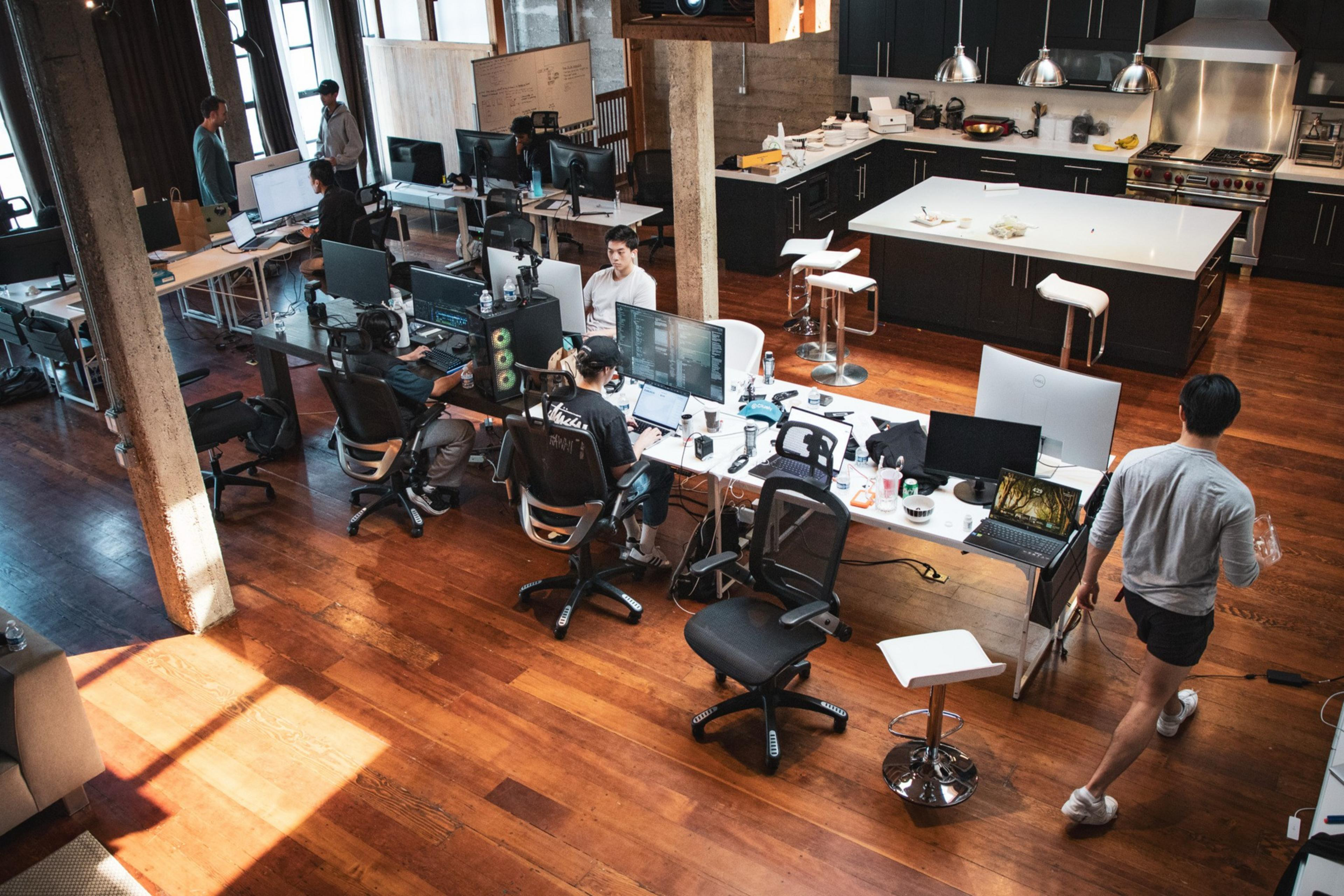 An open-plan office space features people working at desks with multiple computers, a kitchen area in the back, and a wooden floor throughout.