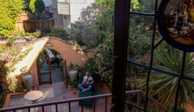 A person sits cross-legged on a green bench in a sunlit garden patio, surrounded by plants, flowers, large pots, an archway, and another person beyond it.