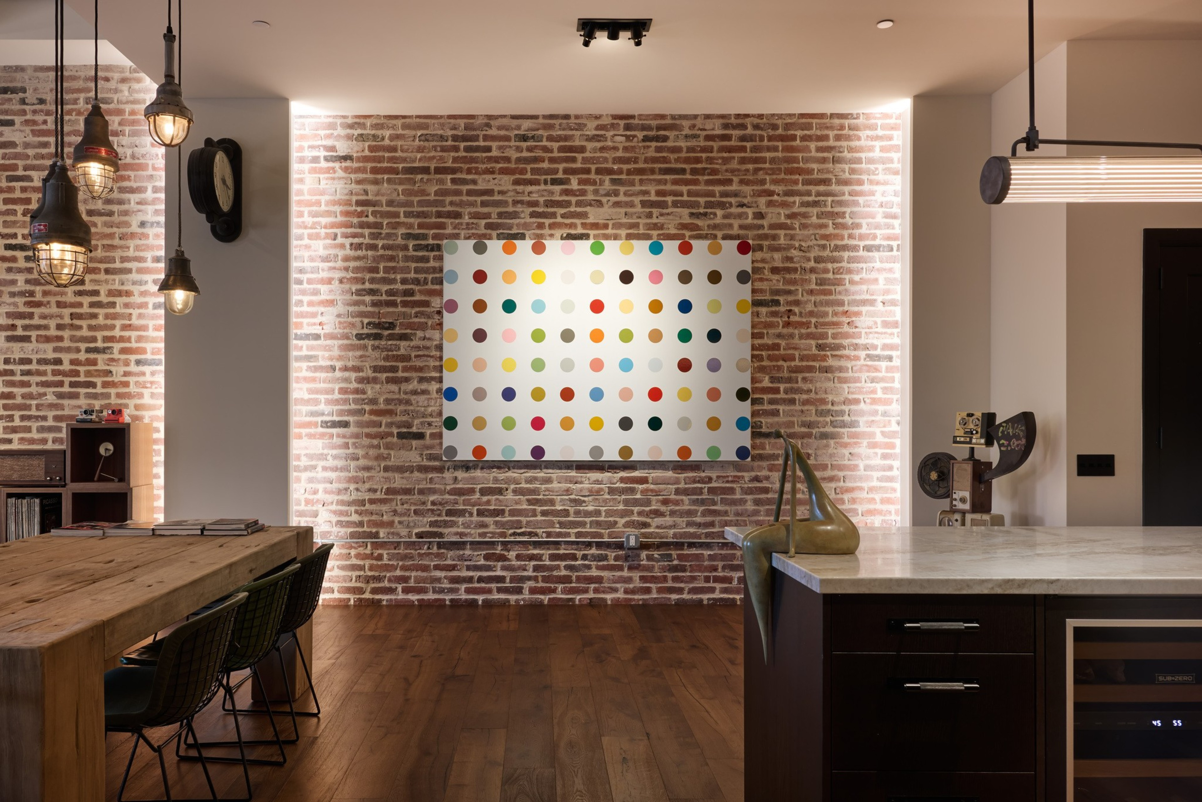 A modern room with exposed brick wall features a colorful polka dot artwork, wooden dining table with chairs, pendant lights, and a kitchen island with decor.