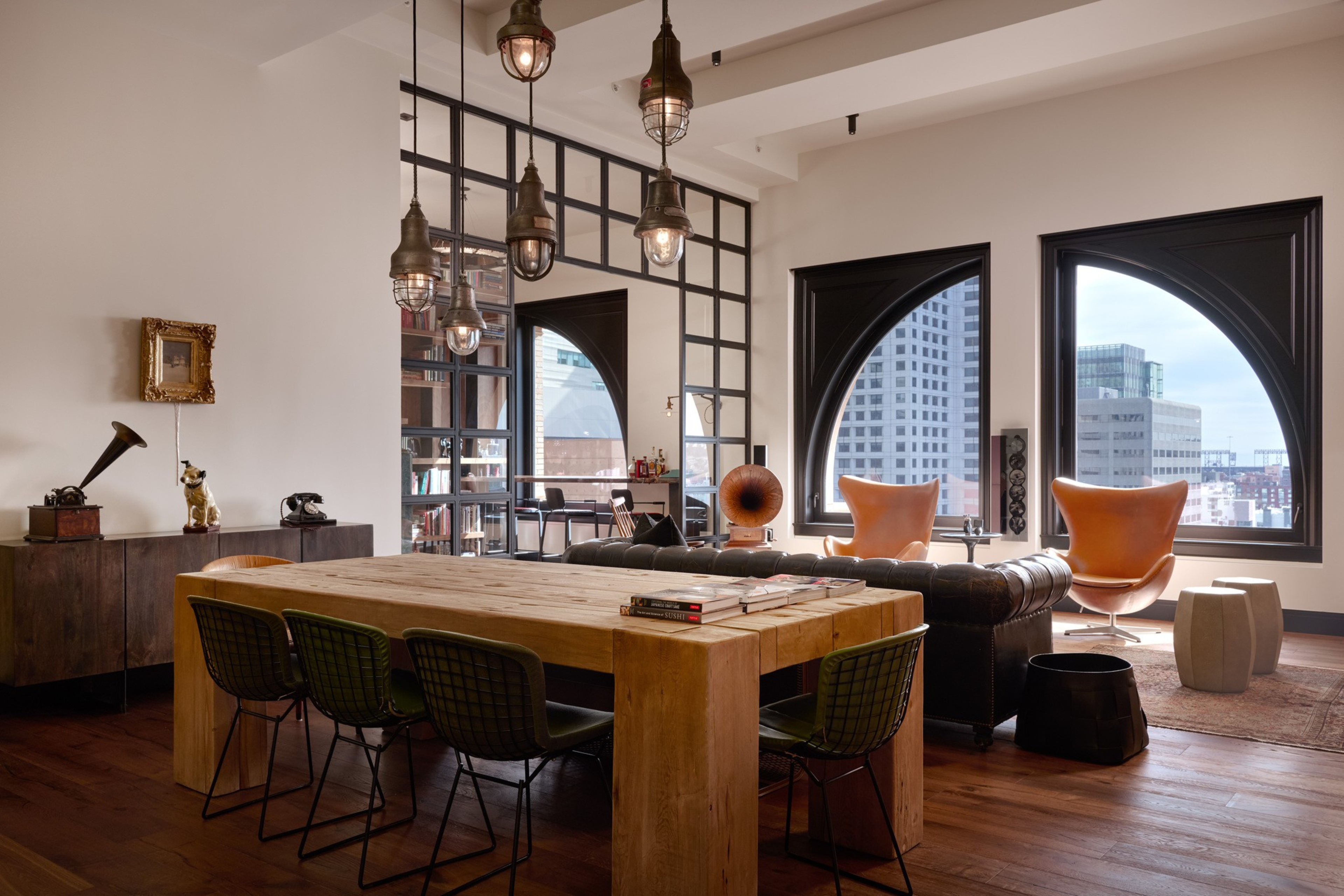 A modern living space features a wooden dining table with black chairs, leather armchairs by large arched windows overlooking a cityscape.