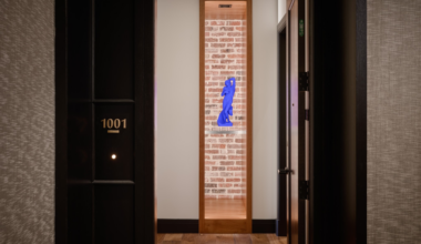 A narrow wooden display niche with a blue modern sculpture set against a brick wall is framed by a beige hallway, with black doors on each side.