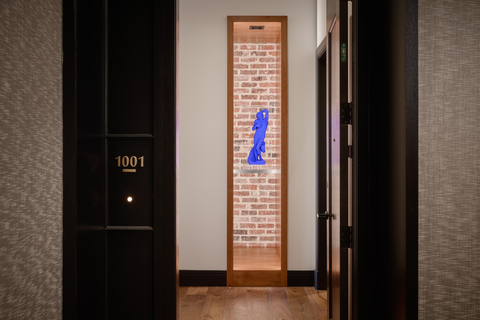 A narrow wooden display niche with a blue modern sculpture set against a brick wall is framed by a beige hallway, with black doors on each side.