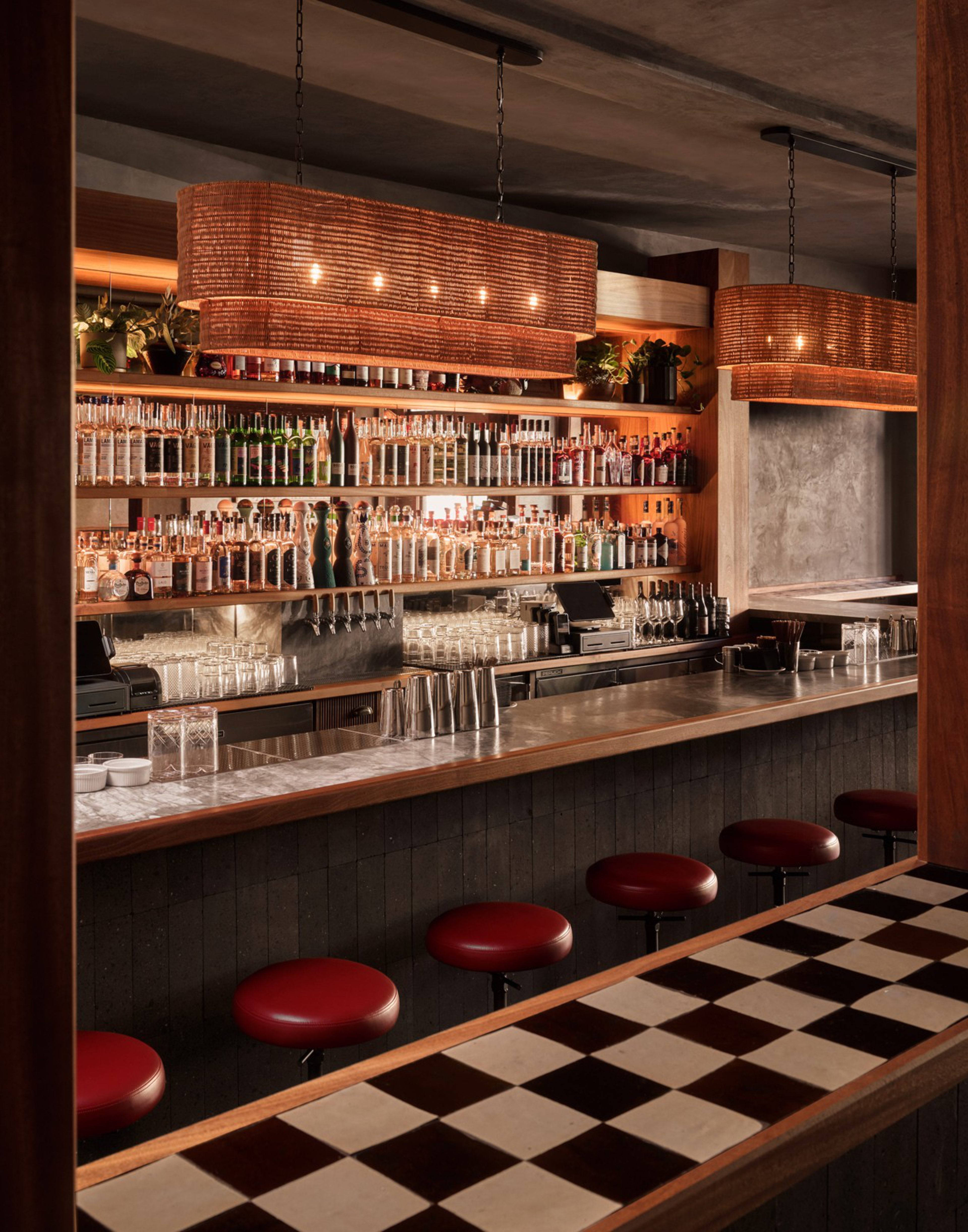 A dimly lit bar with a checkered countertop, red round stools, hanging woven lights, and shelves stocked with various bottles of alcohol.