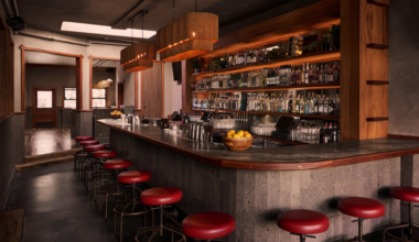 A sleek bar with concrete counters, red cushioned stools, wooden shelves stocked with bottles, and warm hanging wicker light fixtures.