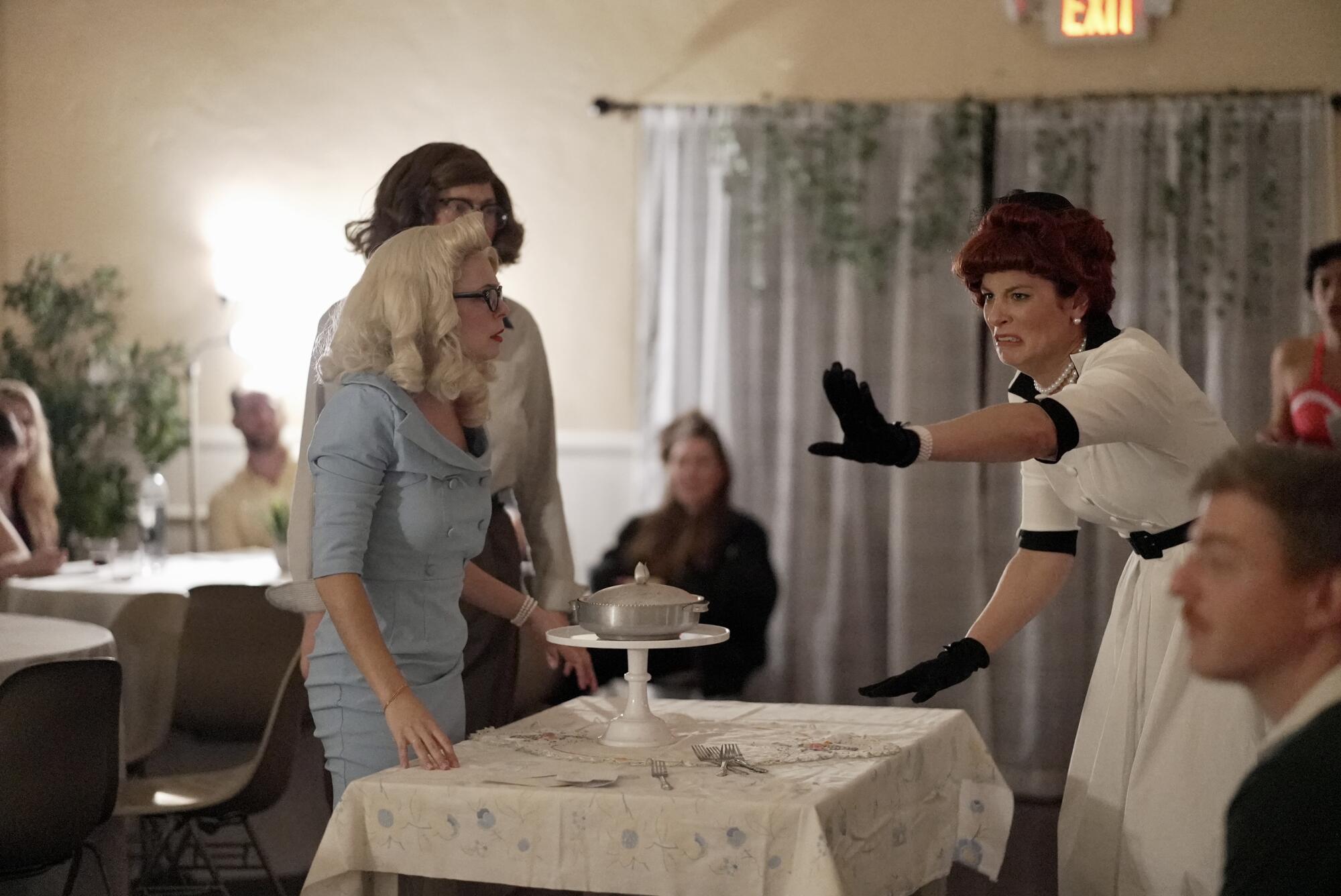 Three actors in 1950s period garb surround a table with breakfast. 
