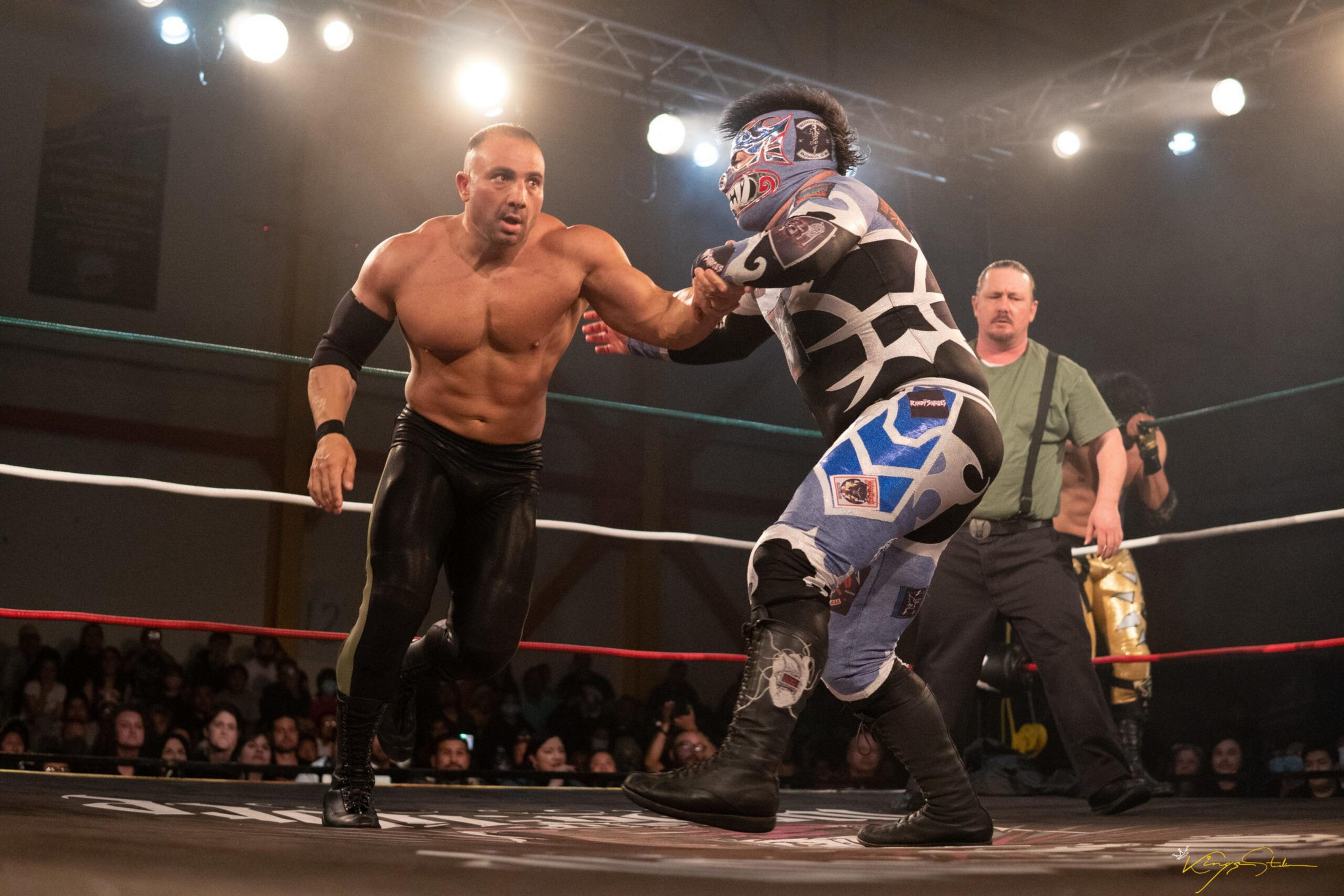 A shirtless wrestler in black tights grips the arm of a masked wrestler in blue and black gear inside a lit wrestling ring with a referee watching.