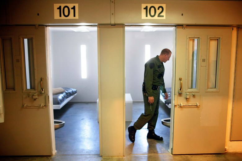 A view of two open prison cells doors inside viewed from the hallway looking in as a prison guard looks around inside the cell on the right. The guard is backlight by light shinning in from a small window towards the back of the cell.