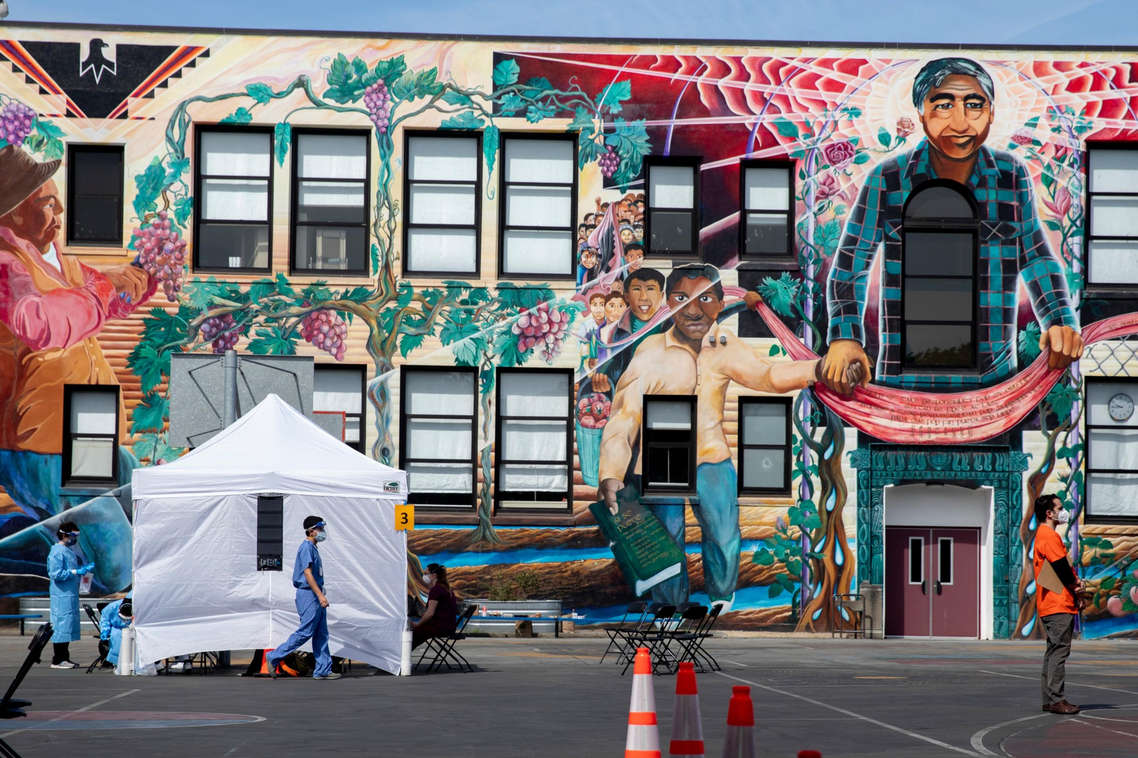 A colorful mural on a building shows people harvesting grapes and holding books, with healthcare workers and a tent set up in front.