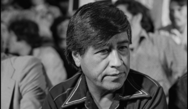 A man with dark hair and a serious expression wears a collared shirt, sitting among a blurred crowd in a black and white setting.