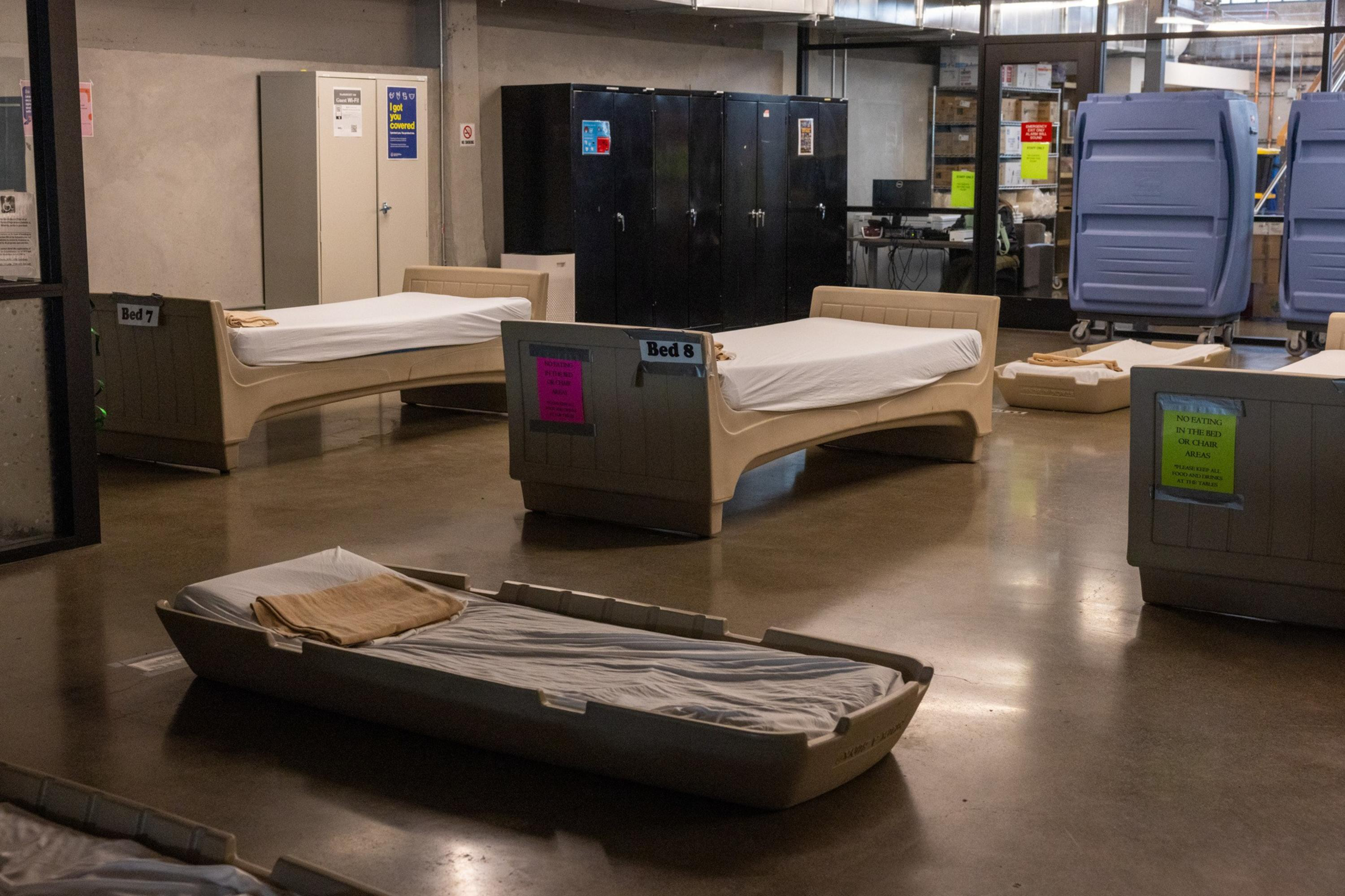 Several plastic beds with thin mattresses and folded blankets are arranged on a concrete floor inside a large, industrial-looking room.
