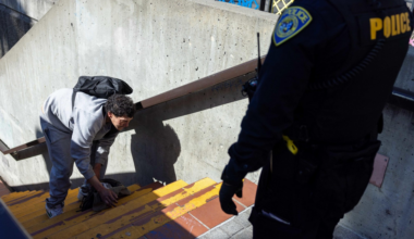 A person in gray sweatpants and a backpack leans down on stairs. A police officer in uniform stands nearby, observing.