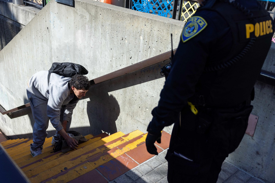 A person in gray sweatpants and a backpack leans down on stairs. A police officer in uniform stands nearby, observing.