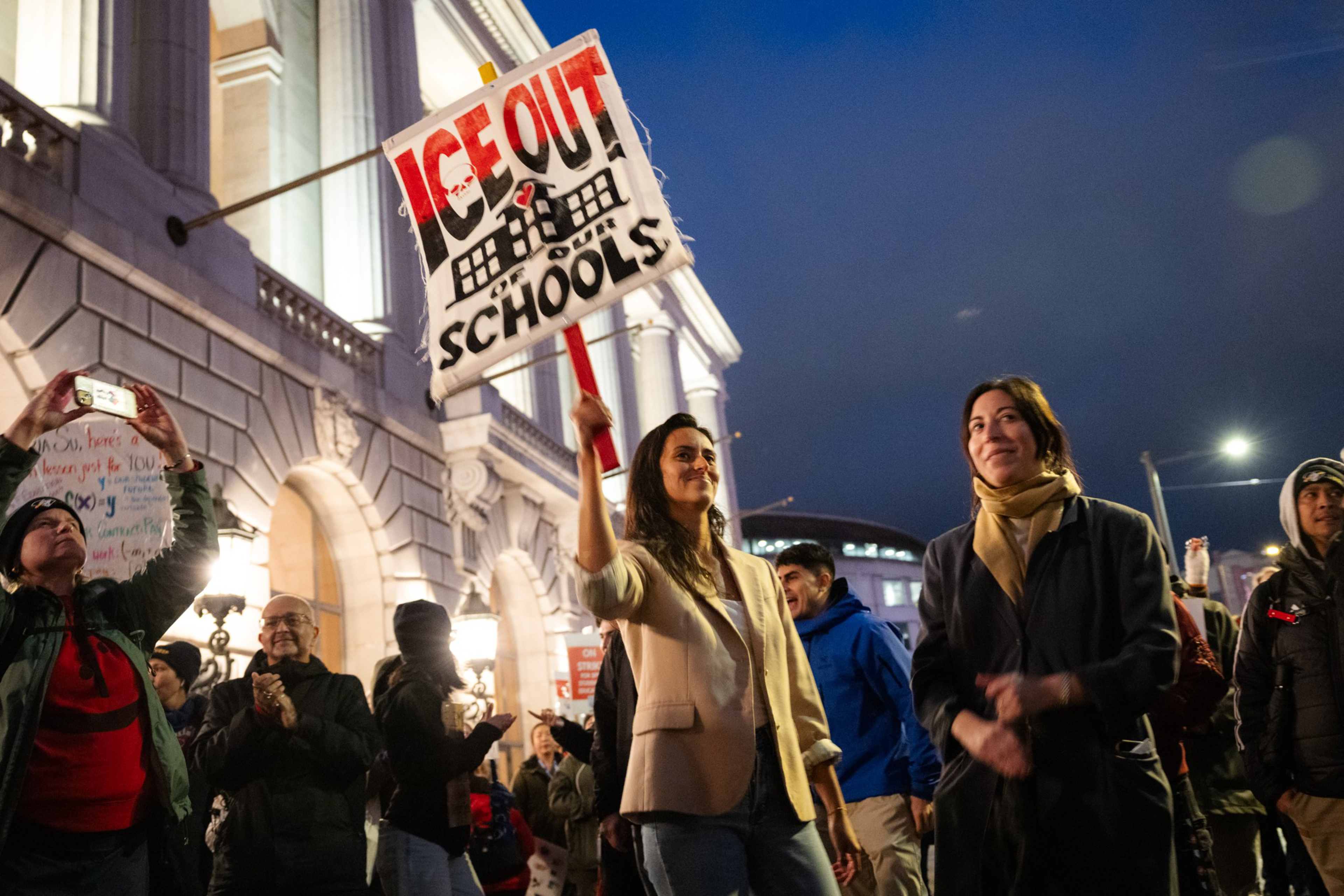 A woman holds a sign that says “ICE OUT OF SCHOOLS” while others around her smile and stand in front of a large, illuminated building at dusk.